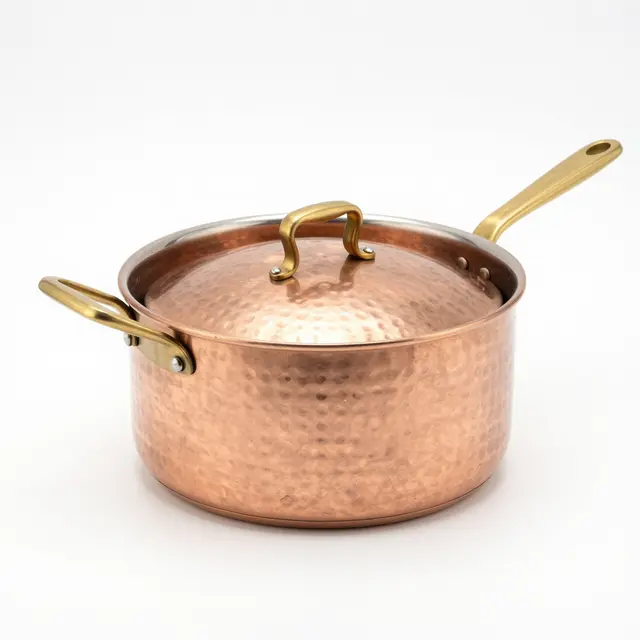 Hammered copper saucepan with brass handles and lid sits isolated on a plain white studio background