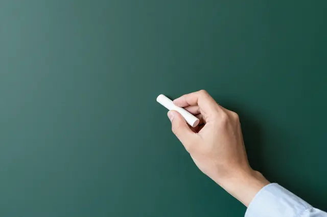 A person's hand holds white chalk poised to write on a blank dark green chalkboard