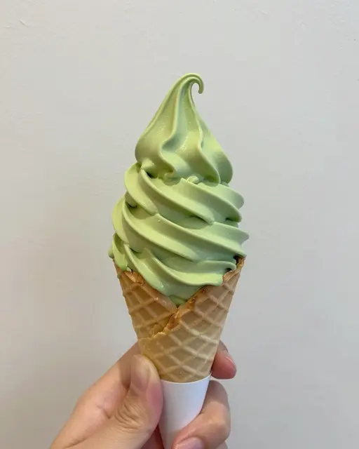 A partial hand holds a waffle cone of green matcha soft serve against a plain white wall