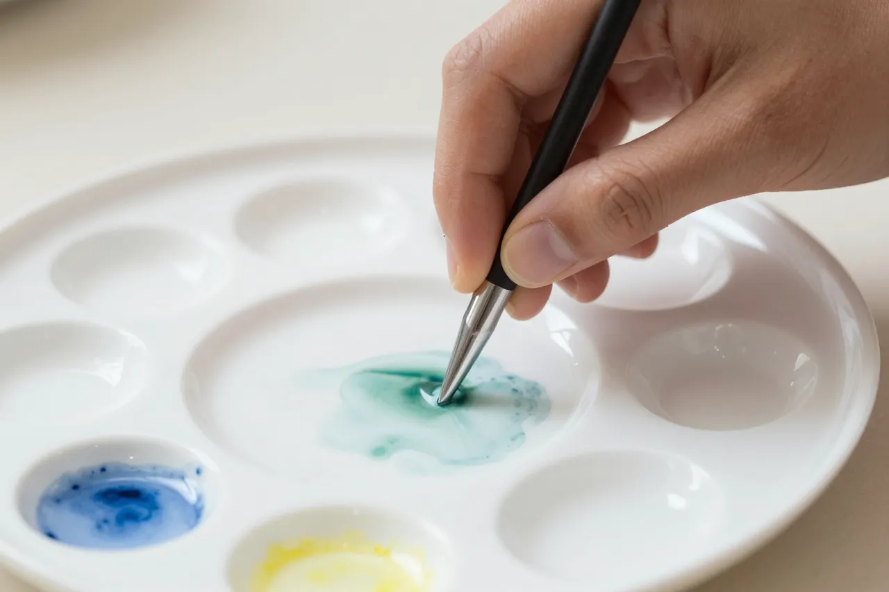 A hand holds a paintbrush to mix green watercolor on a white porcelain artist palette