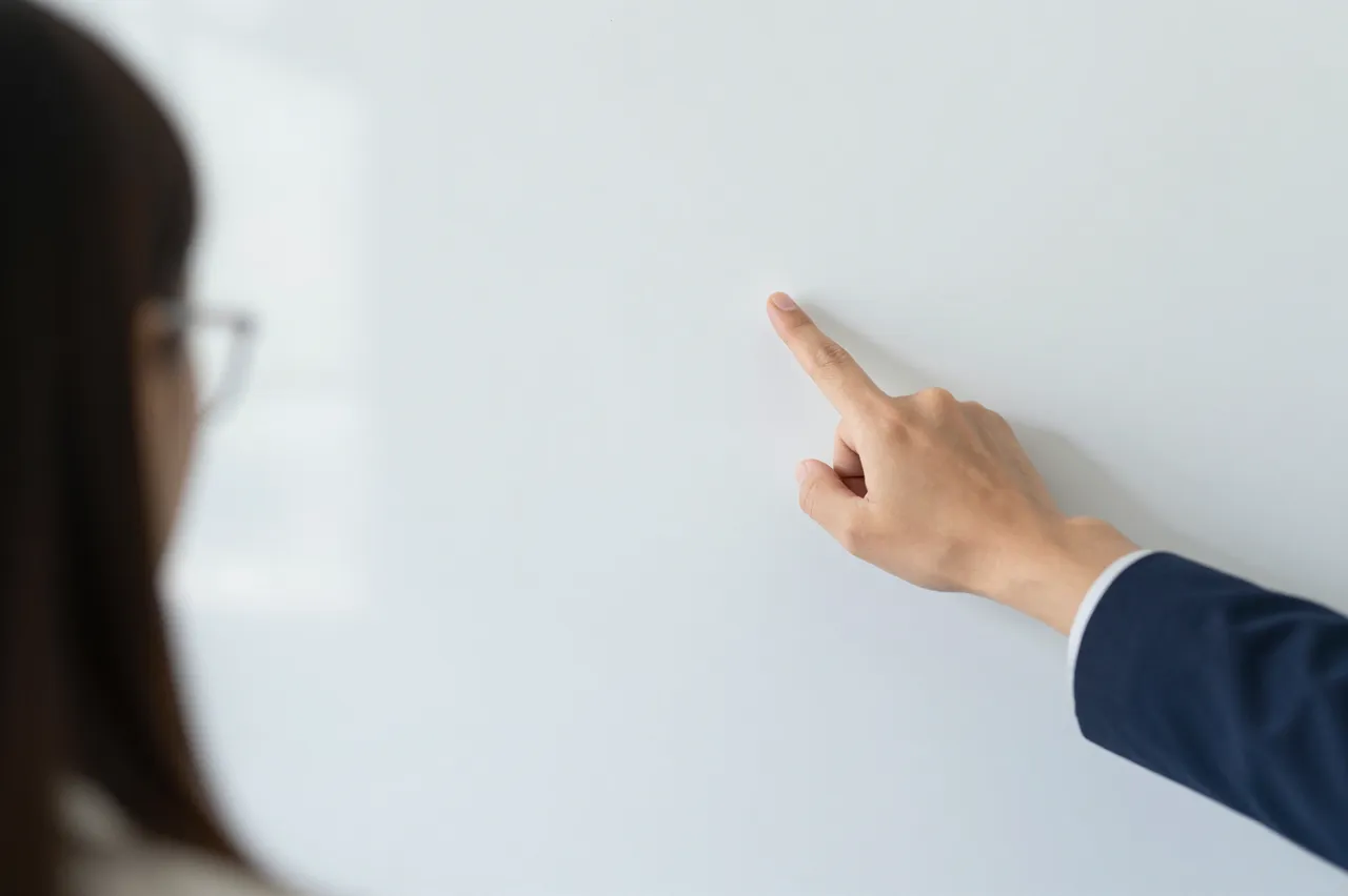 A hand in a navy suit sleeve points at a blank whiteboard, with a blurred woman in glasses observing from the side.