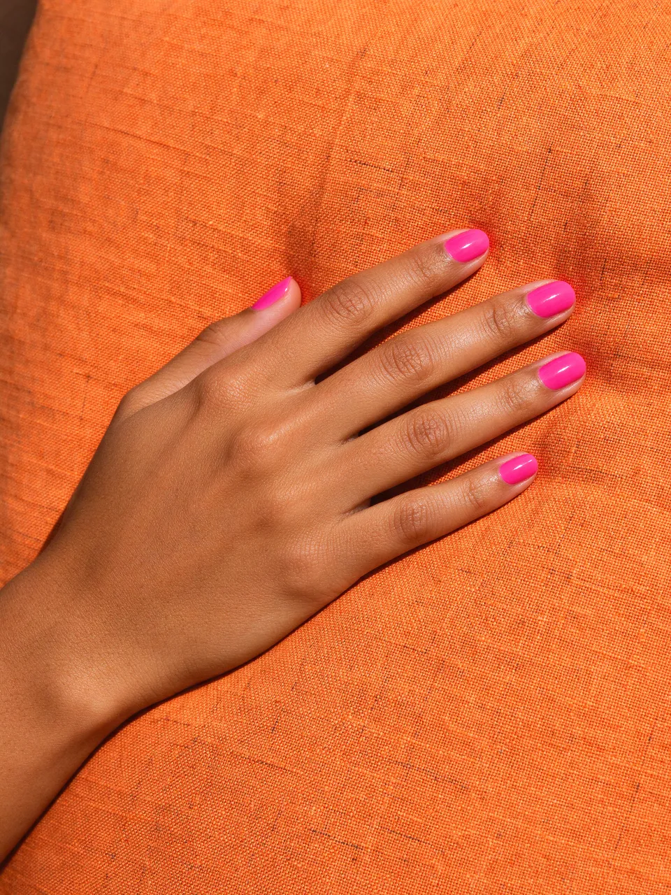A hand with glossy hot pink nail polish rests flat on a textured orange linen cushion in bright light.