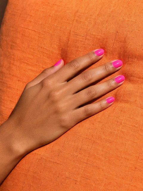 A hand with glossy hot pink nail polish rests flat on a textured orange linen cushion in bright light.