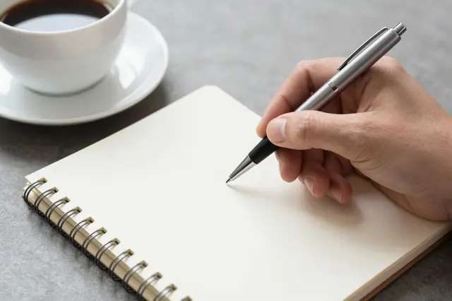 A hand holding a pen hovers over a blank spiral notebook next to a cup of black coffee on a gray tabletop