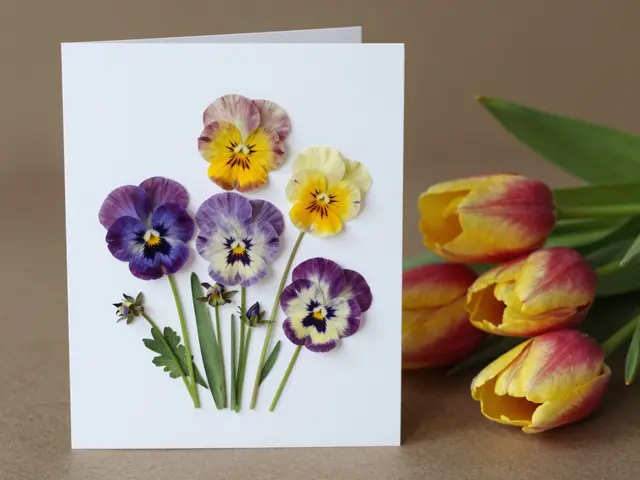Closed handmade card decorated with pressed pansies sits next to red and yellow tulips on a neutral brown surface.