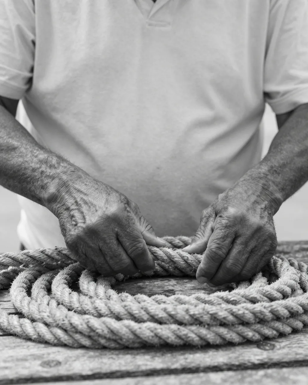 Black and white close-up of an older man's weathered hands coiling thick rope on a wooden dock