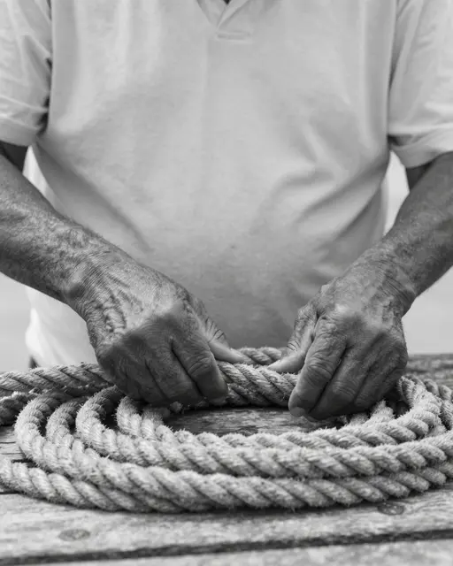Black and white close-up of an older man's weathered hands coiling thick rope on a wooden dock