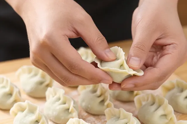 Close-up of a person's hands pleating a handmade dumpling above a wooden board with finished uncooked dumplings.