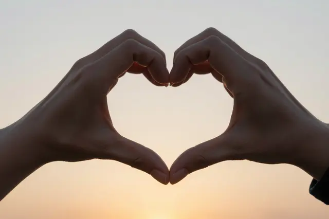 Two silhouetted hands form a heart shape against a soft gradient sunset sky.