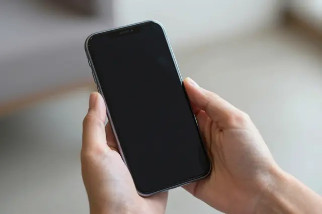 Two hands hold a smartphone with a blank dark screen against a soft blurred indoor background