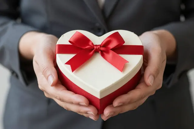 Cupped hands hold a small heart-shaped gift box tied with a red satin bow