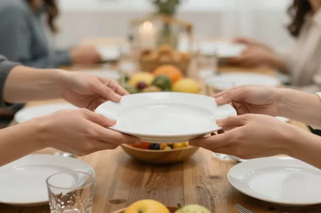 Multiple pairs of hands pass a white plate across a wooden holiday dinner table set with fruit and glassware