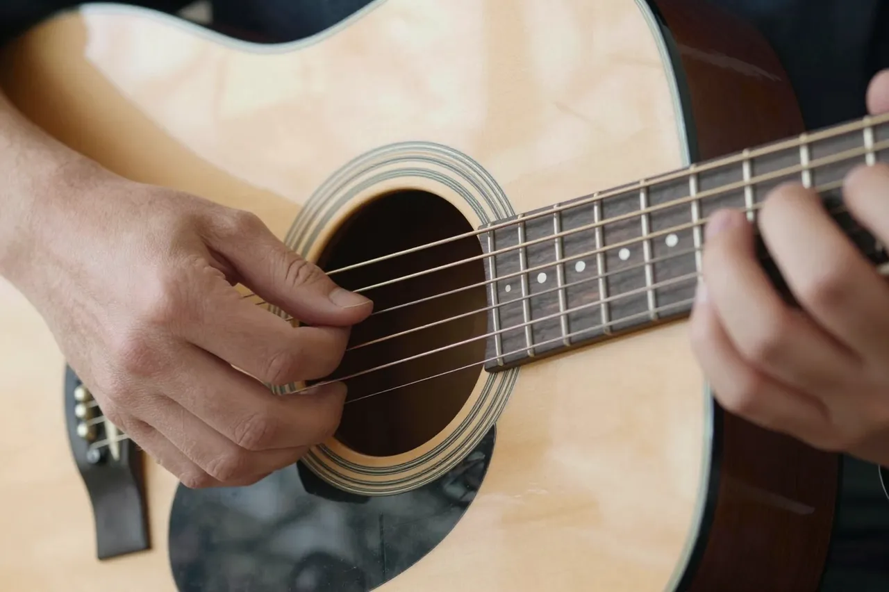 Close-up view of two people's hands fretting and plucking the strings of a natural wood acoustic guitar