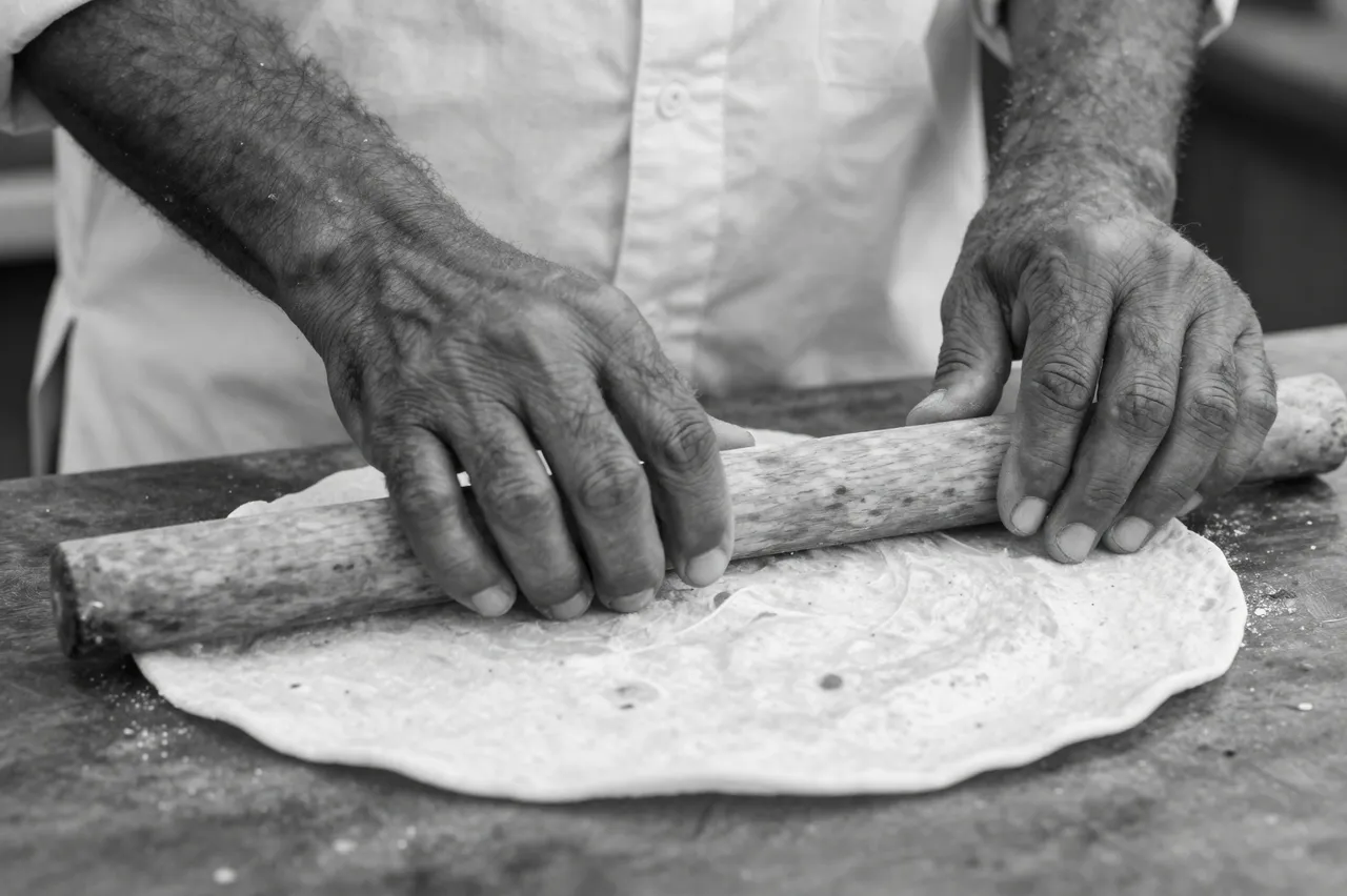Black and white close-up of a man's hands rolling flatbread with a wooden rolling pin on a work surface