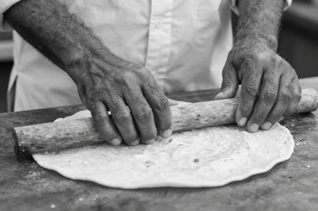 Black and white close-up of a man's hands rolling flatbread with a wooden rolling pin on a work surface