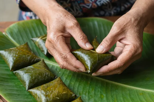 Older Southeast Asian hands fold a square sticky rice cake wrapped in fresh banana leaf atop a large leaf platter.
