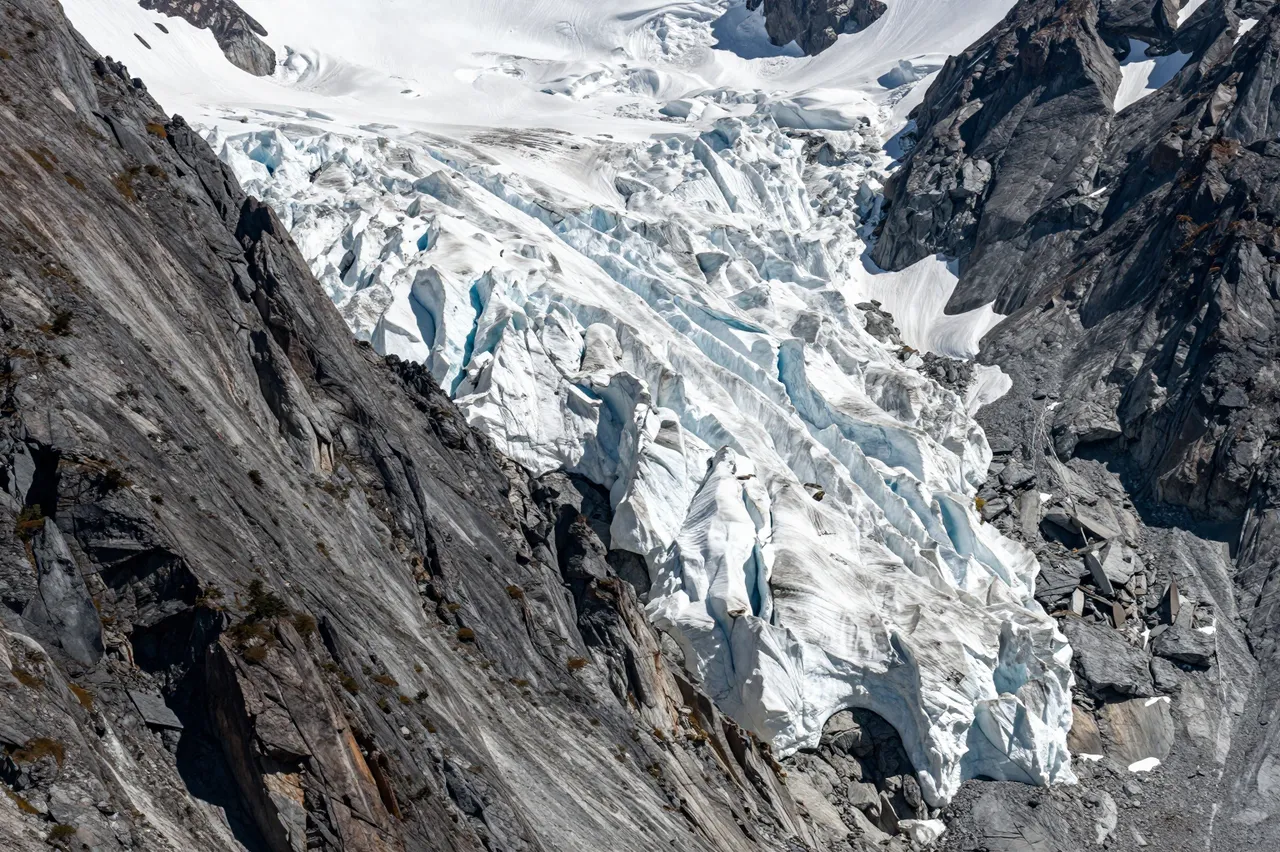 A steep rocky alpine mountain face holds a jagged blue-tinged hanging glacier viewed with a telephoto lens
