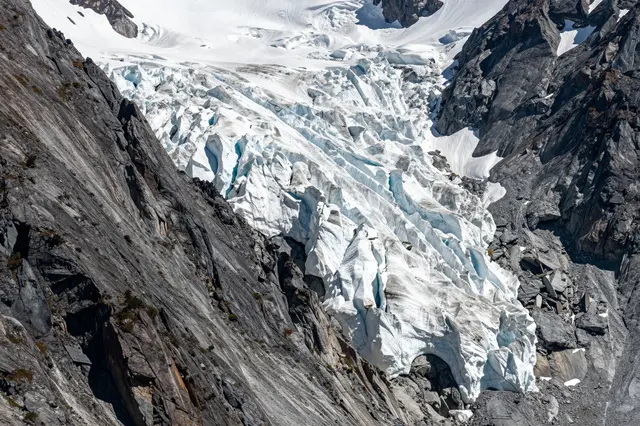 A steep rocky alpine mountain face holds a jagged blue-tinged hanging glacier viewed with a telephoto lens