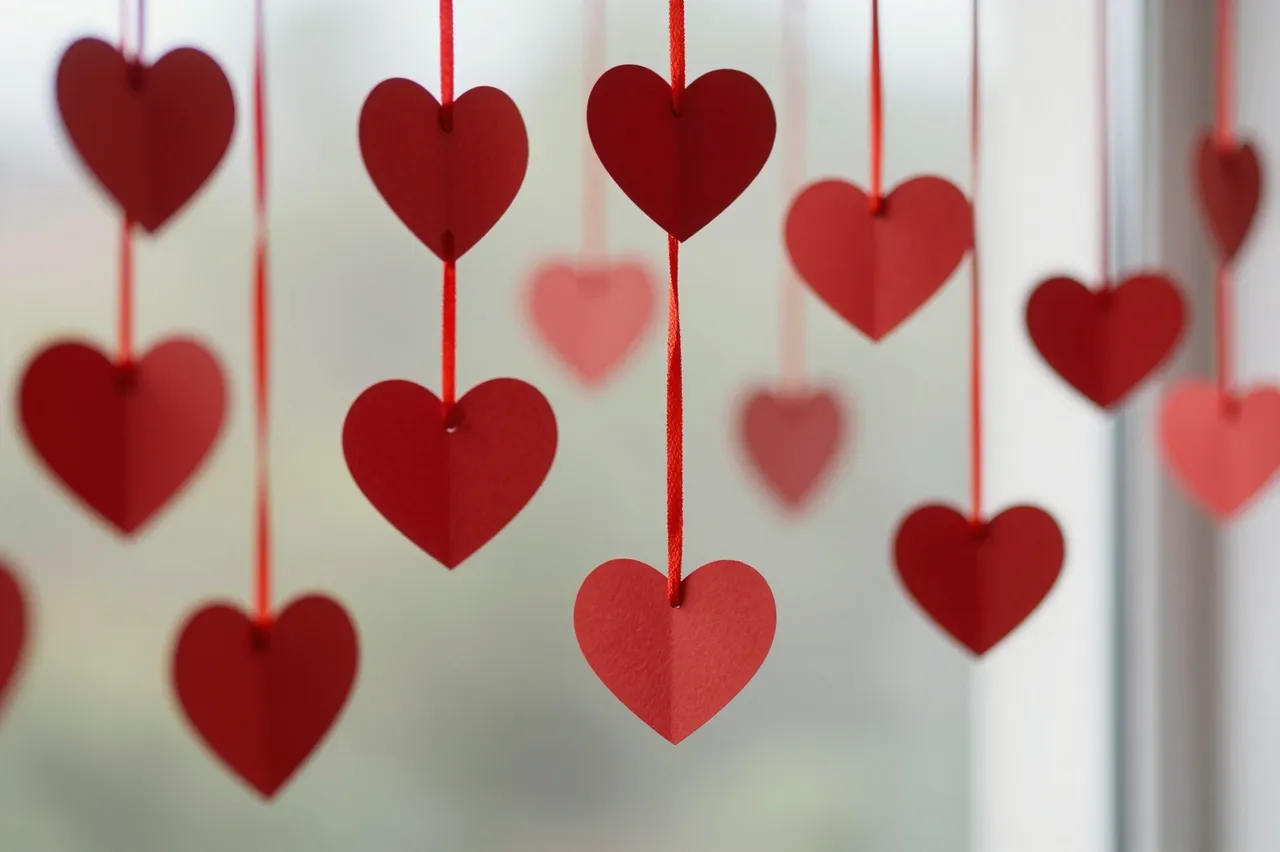 Red paper heart cutouts hang from red strings in front of a blurred bright window background