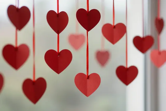 Red paper heart cutouts hang from red strings in front of a blurred bright window background