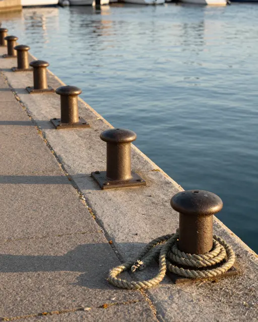 Row of iron mooring bollards on a stone harbor quay, with coiled rope, calm water and distant boats in warm golden hour