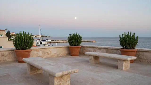 Stone rooftop terrace with benches and potted rosemary overlooks a small coastal harbor under a rising moon