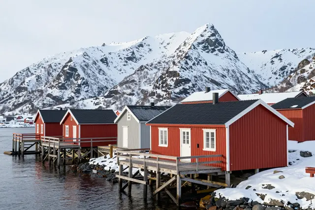 Red and gray wooden boathouses line a cold northern harbor below snow-covered mountain peaks