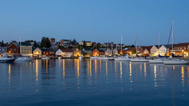 Coastal harbor town with lit waterfront buildings and anchored sailboats reflects in calm water at blue hour