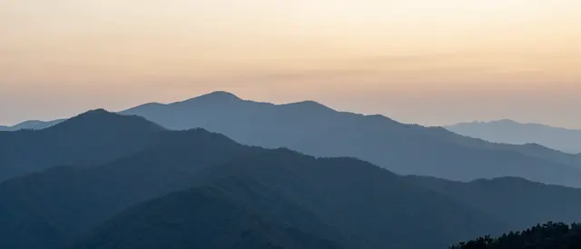 Layered smoky blue mountain ridges fade into soft haze under a warm sunset sky