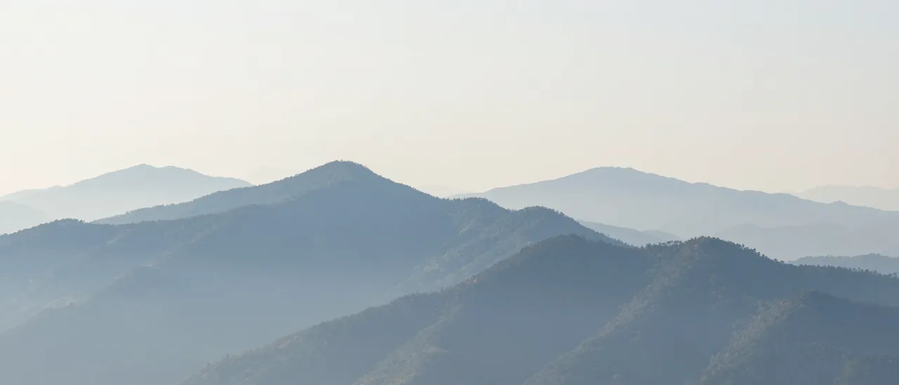 Layered blue mountain ridges fade into soft morning mist beneath a pale open sky