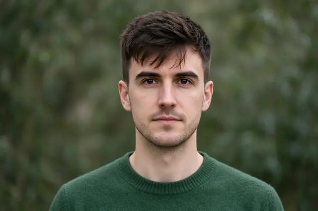 Head and shoulders portrait of a man in a green knit sweater looking at the camera against blurred foliage
