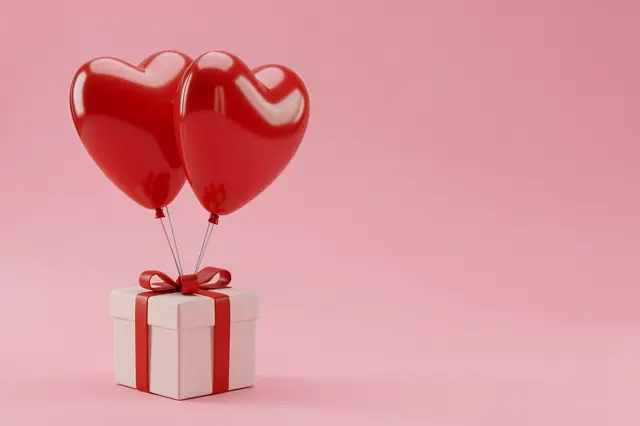 Two glossy red heart balloons tied to a ribboned gift box on a solid pink backdrop with empty right side space