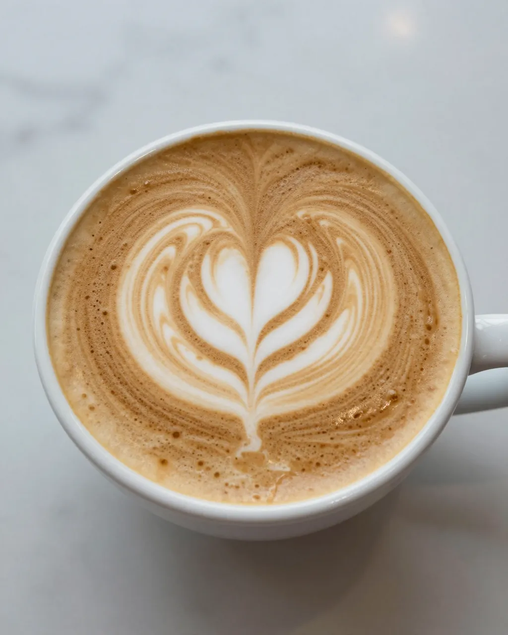 Top-down view of a white coffee cup holding a latte with heart latte art on a light marble table.
