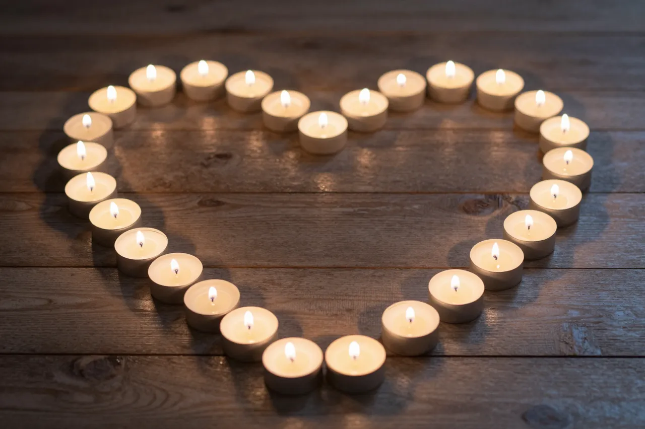 Lit tealight candles arranged in a neat heart shape rest on dark wooden planks at night