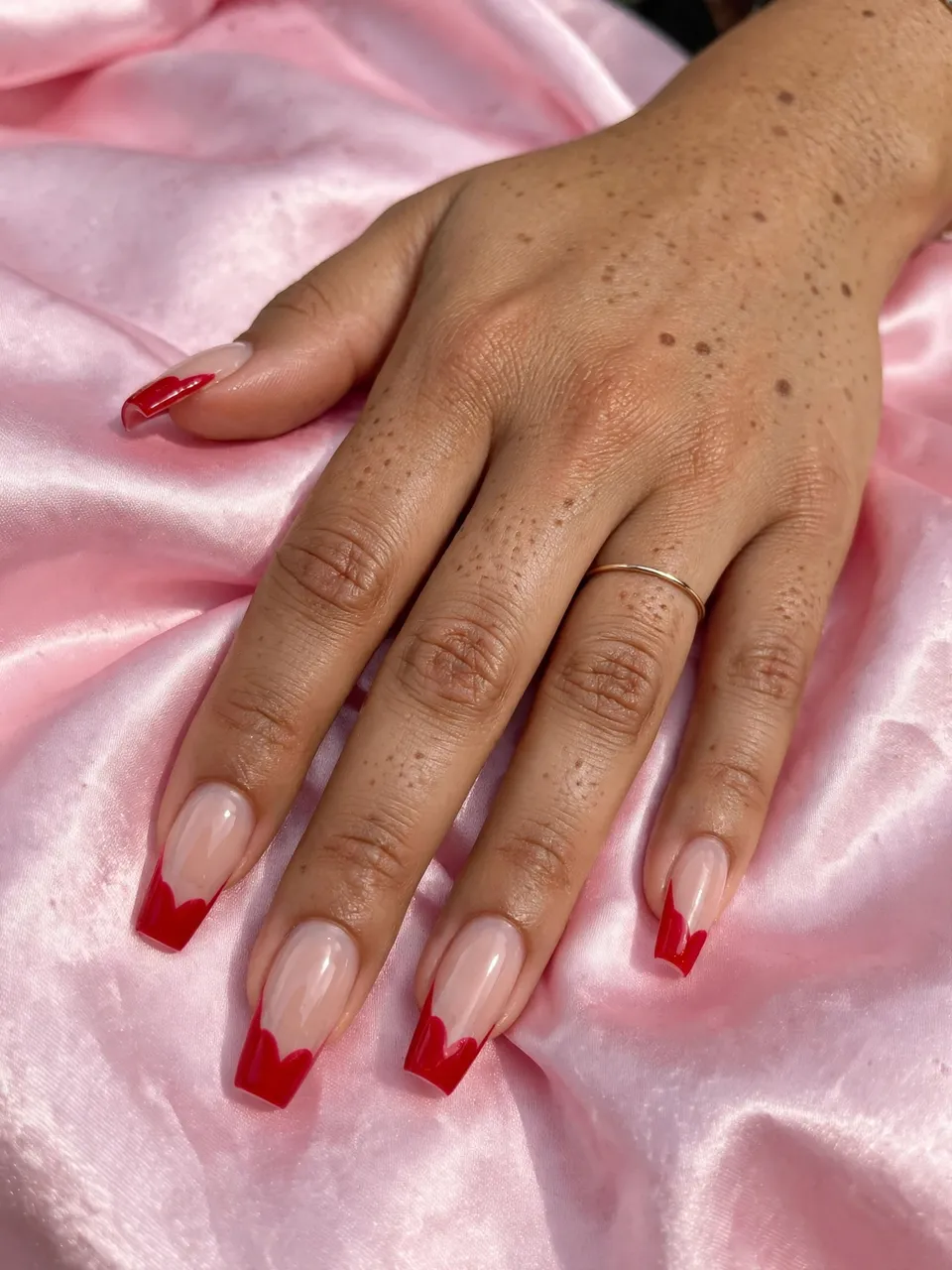 A freckled hand with red heart French tip coffin nails rests on soft pink satin.