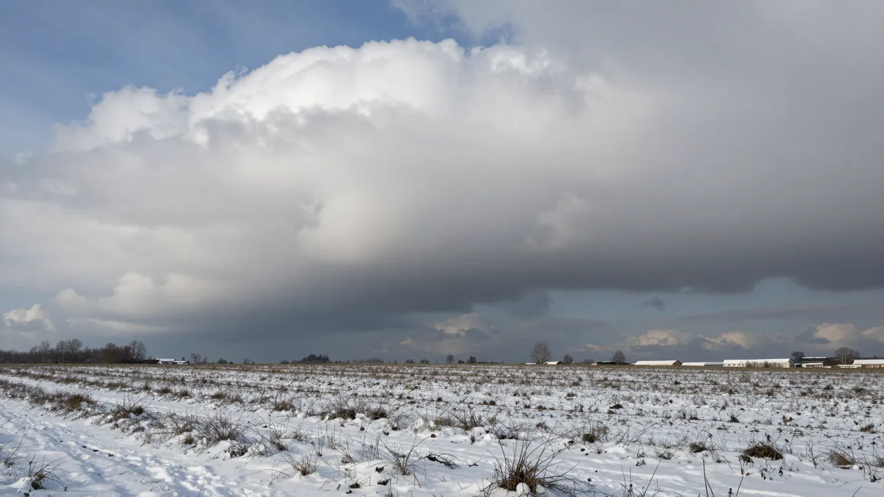 Towering heavy snow clouds loom over a snow-dusted bare winter agricultural field