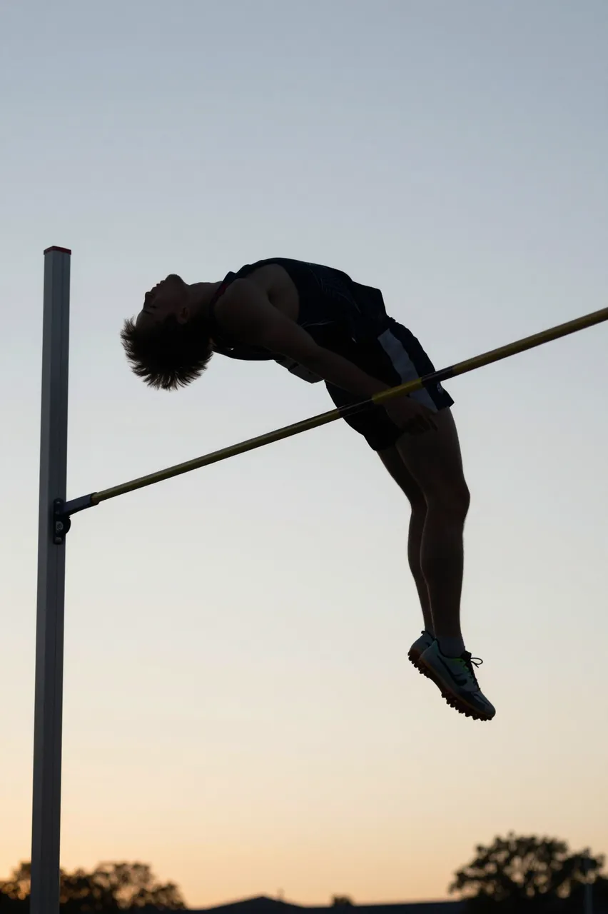A high jumper arches over the high jump bar against a soft gradient evening sky