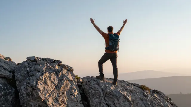 A back view of a hiker standing on rocky mountain summit with both arms raised at sunrise