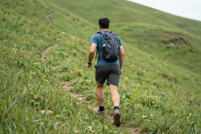 A man seen from behind hikes along a dirt trail up a lush green grassy hill