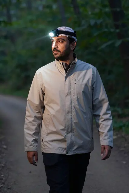 Middle Eastern man walking a dusk forest trail wearing a lit headlamp and light jacket