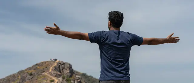Back view of a South Asian man standing on a mountain ridge with arms outstretched under a hazy sky