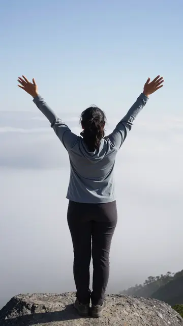 Back view of a woman standing on a rocky peak with arms raised above a sea of clouds