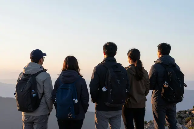 Group of adult hikers stand on a mountain summit facing the rising sun, viewed from behind