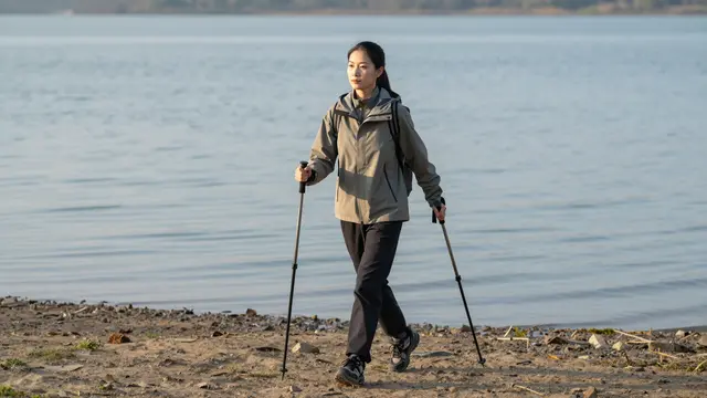 An East Asian woman in hiking gear walks with trekking poles along a calm lake shore at dawn