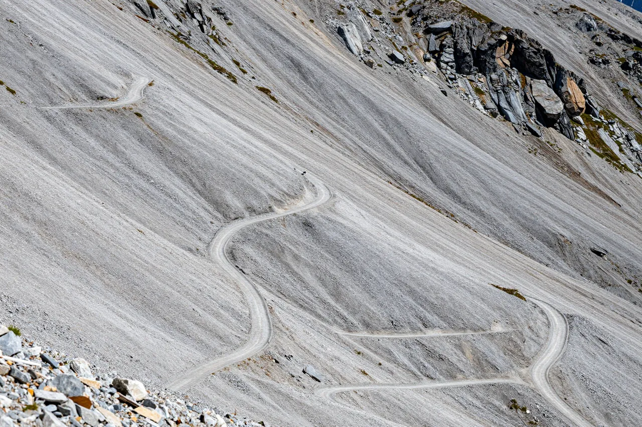 A high-angle view of zigzagging hiking trail switchbacks climbing a gray rocky scree mountain slope