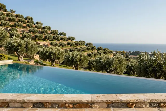 Infinity resort pool with stone edge overlooks terraced olive groves and a distant sea under clear late afternoon sky