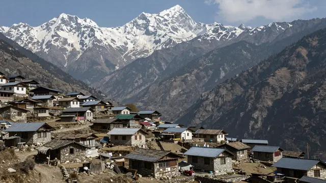Stone village houses sit on a hillside beneath snow-capped Himalayan peaks under a clear blue sky