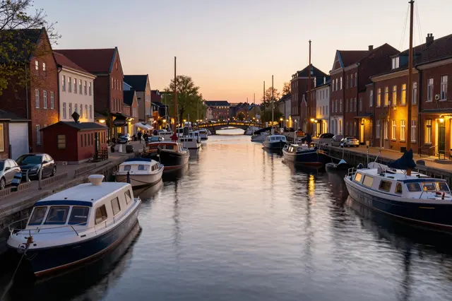 Moored line boats line a quiet historic city canal as warm street lights glow at dusk