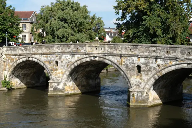 A weathered historic stone arch bridge spans a slow river in a sunlit old town