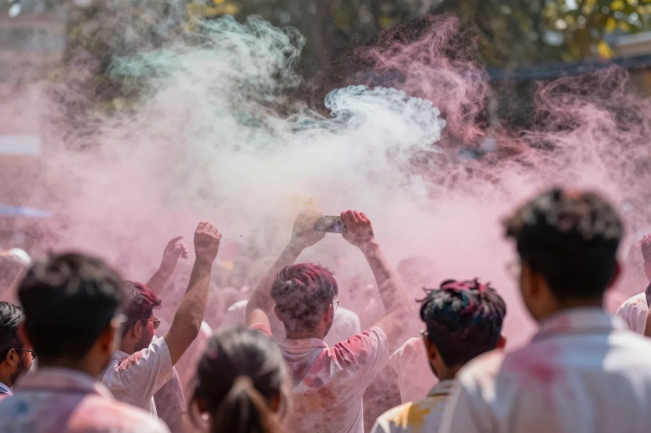 Group of adults viewed from behind amidst swirling colored powder at an outdoor Holi festival